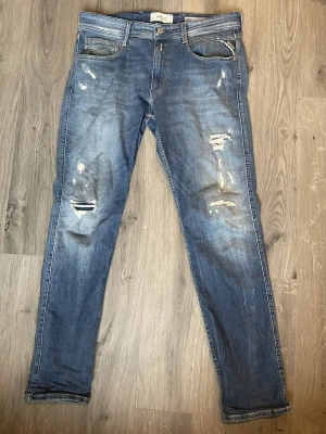 Blå Replay jeans med slitningar, W32 L32 - Säljer ett par blå jeans från Replay i modell Anbass med snygga slitningar och distressed detaljer på benen. Klassisk femficksmodell med raka ben och normal passform. Jeansen har en skön tvättad look och är tillverkade i slitstarkt denimtyg.