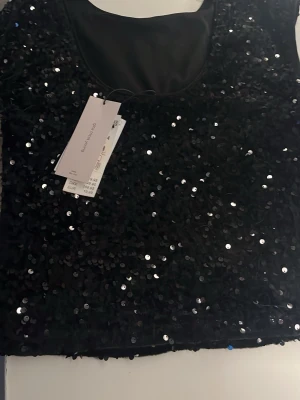 Svart paljettopp från H&M - Säljer en svart glittrig topp från H&M, täckt med paljetter som ger ett riktigt snyggt och festligt intryck. Toppen är ärmlös och har en rund halsringning. Perfekt för dig som vill sticka ut och glänsa lite extra.