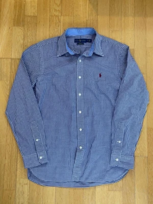 Blårutig skjorta från Ralph Lauren - Snygg blårutig skjorta från Ralph Lauren i slim fit. Skjortan har klassisk krage, vita knappar och den ikoniska röda loggan broderad på bröstet. Tillverkad i 100% bomull för en skön känsla. Perfekt till jeans eller chinos.