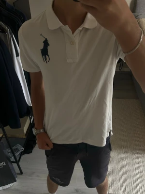 Vit pikétröja från Ralph Lauren - Vit pikétröja från Ralph Lauren med klassisk krage och två knappar framtill. Stor mörkblå logga på bröstet och en stor siffra 3 på ena ärmen. Tillverkad i bomull och har korta ärmar. Perfekt till en avslappnad men ändå stilren look.