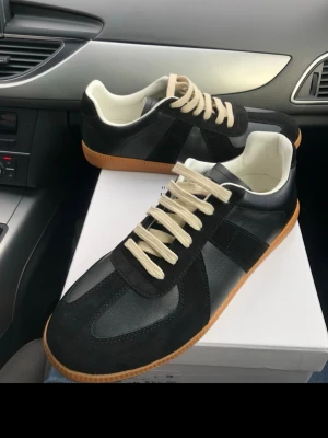 Maison Margiela svarta sneakers i skinn - Maison Margiela sneakers i svart skinn och mocka med beige snörning och vit insida. Snygg kontrast med gummisula i brunt och klassisk design. Perfekta för dig som gillar stilrena och tidlösa sneakers med premiumkänsla.