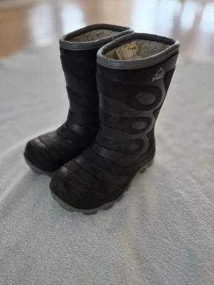 Svarta gummistövlar från McKinley - Säljer ett par svarta vinterstövlar från McKinley i storlek 30. De har grov sula för bra grepp och är fodrade med mjukt syntetmaterial för extra värme. Stövlarna har ett diskret mönster på sidan och är perfekta för kalla dagar.