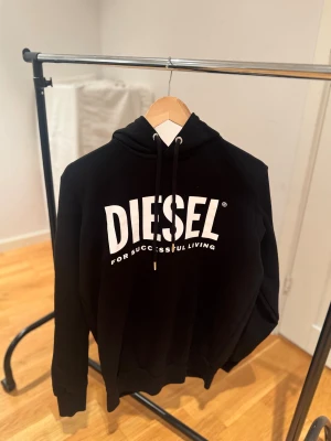 Svart hoodie från Diesel  - Svart hoodie från Diesel i storlek M. Tröjan har huva med snörning och ribbade muddar vid ärmslut och nederkant. Klassisk och stilren. Använts väldigt sparsamt, väldigt bra skick.