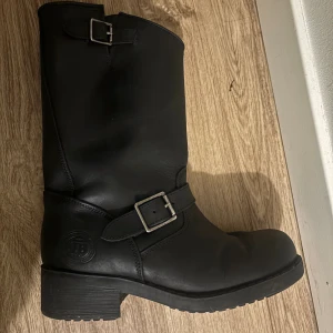 johnny bulls - Säljer ett par svarta boots från JB med grov klack och rund tå. Skorna är tillverkade i skinn och har två silvriga spännen som snygg detalj. Insidan är fodrad med mjukt material för extra värme. Perfekt för kalla dagar och en cool stil.