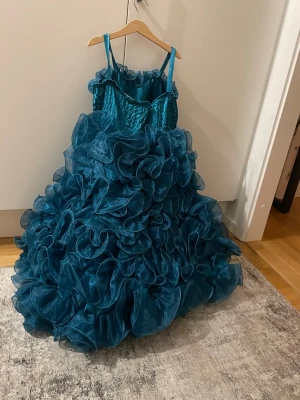 Turkos långklänning med volanger - En riktigt maffig turkos långklänning med tunna axelband och massor av volanger. Livet har ett broderat blommönster och volangkant upptill. Kjolen är superfluffig med lager på lager av transparent tyg som ger en dramatisk look.