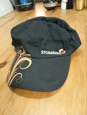 Svart keps från Stora Enso - Svart keps med snyggt guldmönster på skärmen och broderad Stora Enso-logga framtill. Klassisk form med böjd skärm och ventilerande hål upptill. Perfekt för dig som vill ha en unik och stilren keps med cool detalj.