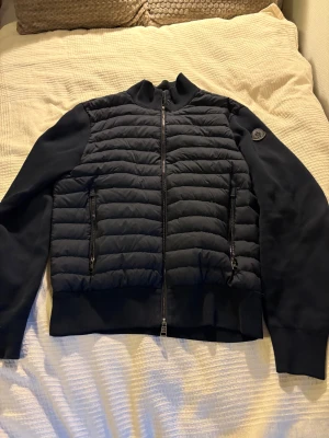 Svart cardigan  från Moncler - Svart cardigan från Moncler med quiltad front och släta ärmar. Jackan har två dragkedjeförsedda fickor, ribbad krage och Moncler-logga på vänster ärm. Materialmix med bomull, perfekt för dig som gillar stilren design.