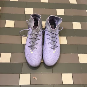 Lila Nike fotbollsskor med snörning - Säljer ett par lila Nike fotbollsskor med högt skaft och snörning. Skorna har en cool grafisk design på sidan och ljusgula detaljer, inklusive den klassiska swoosh-loggan. Utsidan är i syntetmaterial och sulan har dobbar för gräsplan.