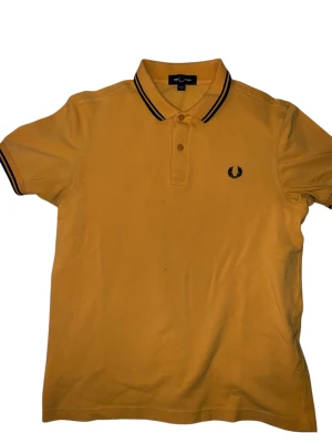 Gul pikétröja Fred Perry - Snygg gul pikétröja från Fred Perry med klassisk svart logga broderad på bröstet. Tröjan har svart och vit rand på krage och ärmslut, samt två knappar framtill. Tillverkad i mjuk bomull och har korta ärmar. Perfekt för dig som gillar sportig och stilren look.