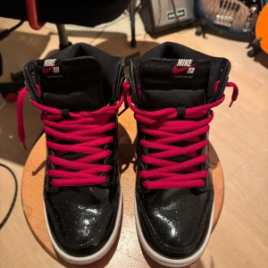 Nike SB Dunk High Pro med röda snören - Nike SB Dunk High bred sneakers i svart med glansig finish och röda snören. Skorna har vit sula, hög modell och extra svarta skosnören medföljer. Ovandelen är i en mix av syntet och textil, med klassisk Nike SB-logga på plösen. Perfekta för dig som gillar streetstyle. No box