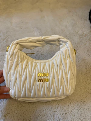 Vit quiltad handväska  - Supersnygg vit handväska med quiltad design och guldiga detaljer. Väskan har ett mjukt, vadderat handtag och guldfärgad logotyp framtill. Perfekt storlek för det viktigaste och ger en lyxig vibe till din outfit.