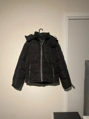 Svart pufferjacka med huva - Svart pufferjacka med huva och dragkedja framtill. Jackan har två fickor med dragkedja och en bröstficka med kardborre. Perfekt för kalla dagar och har en klassisk, lite boxig passform.