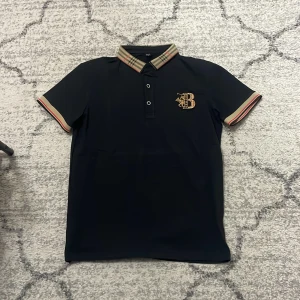 Svart kortärmad skjorta med rutig krage - Svart kortärmad skjorta från Burberry med klassisk rutig krage och ärmslut i beige, rött och svart. Bröstet pryds av ett broderat B-emblem i guld och rött. Skjortan har knappar framtill och är tillverkad i mjukt bomullsmaterial.