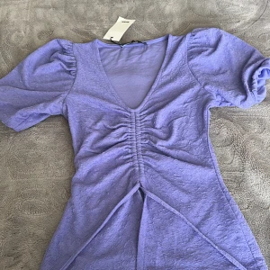 Ljuslila kort klänning från Bik Bok - Supersöt ljuslila kort klänning från Bik Bok med puffärmar, v-ringning och snygg rynkdetalj framtill. Klänningen har en lätt struktur i tyget och markerad midja som ger en feminin siluett. Perfekt för dig som gillar trendiga och färgglada plagg.