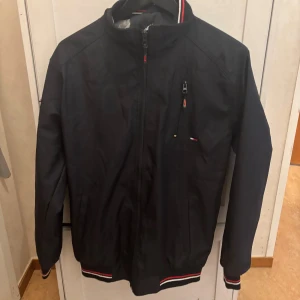 Svart vindjacka Tommy Hilfiger M - Snygg svart vindjacka från Tommy Hilfiger med dragkedja, ribbade muddar och krage med broderad logga. Jackan har detaljer i rött och vitt samt innerfoder med diskret rutmönster. Perfekt för dig som gillar klassisk streetstil.
