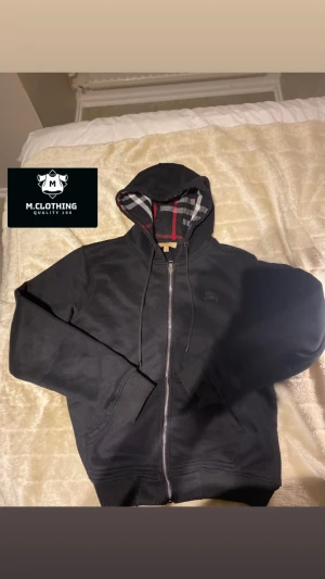 Svart hoodiejacka från Burberry L,M - Svart hoodiejacka från Burberry med dragkedja och klassiskt rutig foder i huvan. Jackan har snörning i huvan, ribbade muddar och två fickor framtill. Perfekt för dig som vill ha en stilren och lyxig streetwear-look.