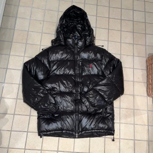 Svart dunjacka från Polo Ralph Lauren - Svart, glansig dunjacka från Polo Ralph Lauren med huva och dragkedja framtill. Jackan har en klassisk pufferstil och den ikoniska röda Polo-loggan broderad på bröstet. Perfekt för kalla dagar och riktigt snygg till streetwear.