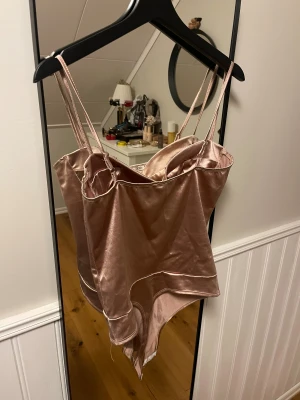 Satin body - Säljer en champagnefärgad  från NA-KD i glansig satin med smala axelband och markerade kupor. Bodyn har en figurnära passform och snygga sömdetaljer, perfekt för fest!
