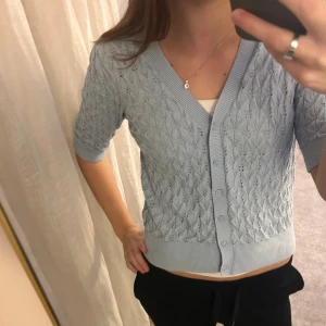 Ljusblå stickad kofta med knappar - Säljer en ljusblå stickad kofta med v-ringning och fina detaljer i mönstret. Koftan har trekvartsärm och stängs med matchande knappar framtill. Ribbad kant vid ärmslut och nederkant ger en snygg finish. Perfekt för dig som gillar mjuka pasteller och söt stil.