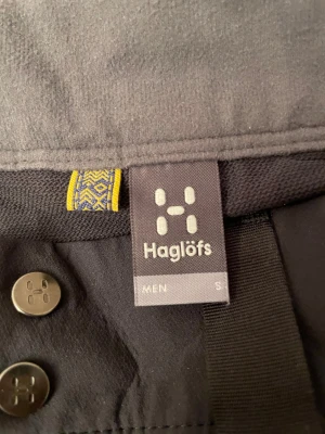 Svarta täckbyxor från Haglöfs herr S - Svarta täckbyxor från Haglöfs i herrmodell, storlek S. Byxorna har två dragkedjeförsedda fickor framtill, bälteshällor och tryckknappar i midjan. Materialet är slitstarkt syntet, perfekt för utomhusaktiviteter under kyliga dagar.