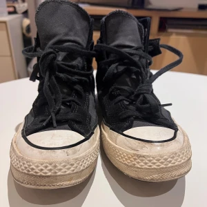 Svarta Converse Chuck Taylor High - Säljer ett par svarta Converse Chuck Taylor high-top sneakers med klassisk vit gummisula och rund tå. Skorna har snörning och är tillverkade i textil med detaljer i mocka. Perfekt för dig som gillar retrostil och vill ha ett par ikoniska sneakers.