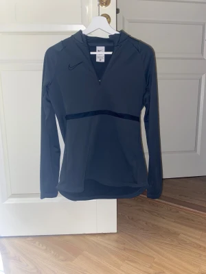 Grå Nike träningströja - Nike Dri-Fit långärmad träningströja i mörkgrå med halv dragkedja framtill och diskret logga på bröstet. Tröjan har en sportig design med markerad söm över magen och är tillverkad i ett lätt syntetmaterial som andas. Perfekt för aktiva dagar och gymmet.