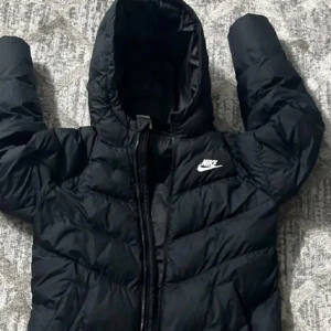 Svart pufferjacka från Nike med huva - Svart pufferjacka från Nike med vit logga på bröstet. Jackan har huva, långa ärmar och dragkedja framtill. Den är quiltad och har en clean, sportig look som passar perfekt när det är kallt ute. Materialet är syntet och jackan är normal i passningen.