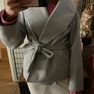 Grå kappa med bälte - Skitsnygg grå kappa i omlottmodell med bred krage och knytbälte i midjan. Jackan har en stilren look och är tillverkad i ett mjukt ullblandat material. Perfekt för dig som gillar en clean och trendig stil.