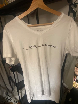Vit t-shirt med tryck från Calvin Klein - Snygg vit t-shirt från Calvin Klein Jeans med v-ringad hals och svart texttryck på framsidan: 'I move ___ in #mycalvins'. Modellen är klassisk med korta ärmar och mjukt bomullsmaterial som känns skönt mot huden. Perfekt för en chill och stilren look.