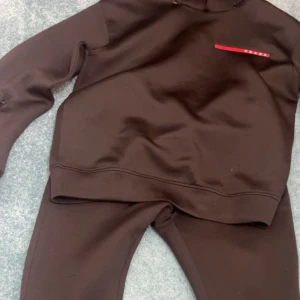 Svart sweatshirt från Prada - Svart sweatshirt från Prada med diskret röd logodetalj på bröstet. Tröjan har en klassisk, avslappnad passform och är tillverkad i 100% bomull för skön komfort. Perfekt för dig som gillar stilrena designer och exklusiva märken.