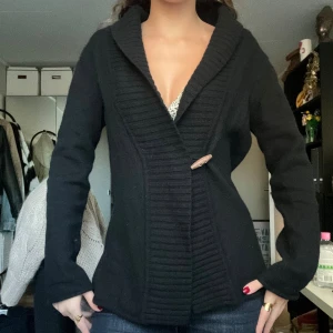 Svart kofta från Boomerang - Svart kofta från Boomerang. Den har en bred ribbad krage och stängs med en träknapp framtill. Materialet är mjukt och stickat, perfekt för kyliga dagar. Mer som S/M än L