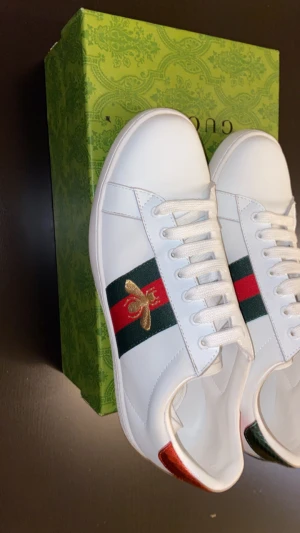 Gucci Ace sneakers med metallic detaljer - Gucci Ace sneakers i vitt skinn med klassiska gröna och röda ränder på sidan. Baksidan har metallicröd och metallicgrön häl med Gucci-logga, och insidan är i mjukt vitt skinn. Snygga snörningar och platt sula. Kommer med originalkartong.