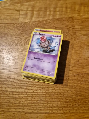 Pokémonkort - lila  - En samling Pokémonkort med Spoink överst. Perfekt för dig som gillar att samla, byta eller spela Pokémon TCG! Färgglada kort med coola illustrationer och attacker. Passar både nybörjare och erfarna samlare som vill utöka sin kortlek.