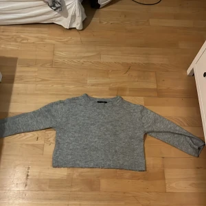 Grå croppad stickad tröja 157 - Grå croppad stickad tröja från 157 med rund hals och långa ärmar. Tröjan har ribbade muddar vid hals och nederkant, vilket ger en chill och enkel vibe. Perfekt för dig som gillar en loose och avslappnad stil. OBS! tröjan är lite sönder men det syns inte på!