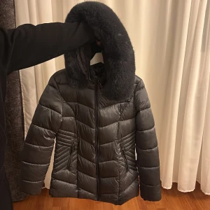 Mörkgrå pufferjacka med fluffig huva - Snygg mörkgrå pufferjacka med stor fluffig huva i fuskpäls. Jackan har dragkedja framtill, två sidofickor med dragkedja och quiltad design. Perfekt för kalla dagar och har en riktigt mysig vibe. Passformen är normal och jackan är långärmad.