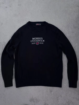 Svart stickad tröja Morris Stockholm - Svart stickad tröja från Morris Stockholm med vit broderad logga och emblem på bröstet. Rund halsringning och långa ärmar med ribbade muddar. Perfekt för dig som gillar klassisk stil med en modern touch.