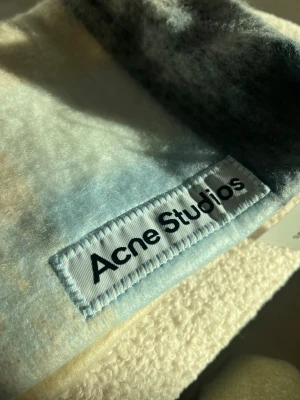 Rutig halsduk Acne Studios i ull - Snygg och fluffig halsduk från Acne Studios med rutigt mönster i ljusblått, beige och grått. Tillverkad i Italien av mjuk ull, perfekt för att ge din outfit en cool touch. Acne Studios-logga är tydligt synlig på etiketten.