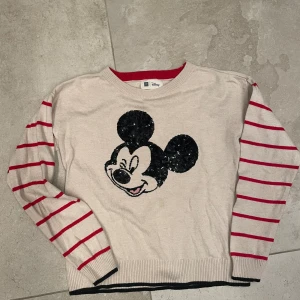Beige stickad tröja med Mickey Mouse GAP - Beige stickad tröja från Gap x Disney med svart paljettbroderad Mickey Mouse på bröstet. Ärmarna är långärmade och har röda ränder, samt ribbade muddar med svarta detaljer. Rund hals med röd ribbad kant. Perfekt för dig som gillar Disney och vill ha en unik look.