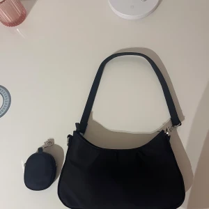 Svart axelväska med mini-plånbok - Snygg svart axelväska i syntetmaterial med stilren, rundad form och silverfärgade metalldetaljer. Väskan har dragkedja upptill och flera fack inuti. Medföljer en matchande mini-plånbok med dragkedja och karbinhake som kan fästas på väskan. Perfekt för att hålla koll på småsaker. Bara att skriva om ni har frågor!!❤️