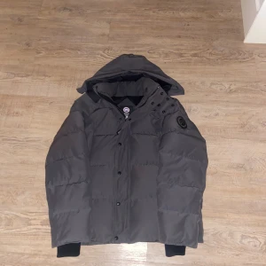 grå wyndham  från Canada Goose - Säljer en grå wyndham  från Canada Goose med avtagbar huva och diskret logga på ärmen. Jackan är tjock och varm, har dragkedja och knappar framtill samt muddar vid ärmsluten. Perfekt för kalla vinterdagar och riktigt snygg streetstil. 