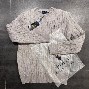 Grå Polo Ralph Lauren - Stickad tröja i ljusgrått från Polo Ralph Lauren med klassisk rund hals och broderad logga på bröstet. Tröjan har ribbade muddar vid ärmslut och nederkant, samt långärmad passform. Perfekt för dig som gillar stilren och tidlös design.