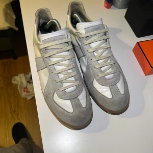 Maison Margiela Gats Vita - Maison Margiela Gats sneakers i vit skinn med ljusgrå mockadetaljer och bruna gummisulor. Klassisk rund tå, snörning och retroinspirerad design. Perfekt för dig som gillar minimalistisk stil och snygga detaljer. Kvitto och dusstbag finns med. 