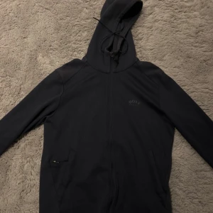 Mörkblå hoodie från BOSS med dragkedja - Mörkblå hoodie från BOSS med hel dragkedja och huva med snörning. Tröjan har två fickor framtill och diskret BOSS-logga på bröstet. Materialet är mjukt och känns skönt mot huden, perfekt för chill eller när du vill ha en clean look.