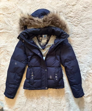 Mörkblå pufferjacka från Luhta - Säljer en mörkblå pufferjacka från Luhta med stor huva och snygg fuskpäls runt kanten. Jackan har dragkedja, knappar och praktiska fickor framtill. Perfekt för kalla dagar med sin vadderade design och coola detaljer.