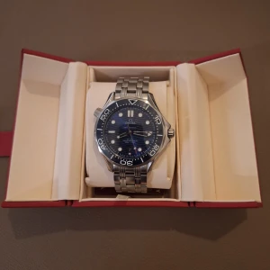 Omega Seamaster blå/silver klocka - Säljer en Omega Seamaster 2018 modell med blå urtavla och vågmönster, silverfärgat armband i rostfritt stål och roterande bezel. Klockan har datumvisning och transparent baksida som visar urverket. Kommer i original ask med certifikat.
