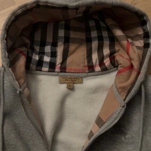 Grå Burberry hoodie med rutig huva - Snygg grå hoodie från Burberry med klassisk rutig insida i huvan och broderad logga på bröstet. Tröjan har dragkedja, snörning i huvan och två fickor framtill. Perfekt för dig som gillar stilrena detaljer och ikoniska mönster. Den är xl men sitter absolut som L