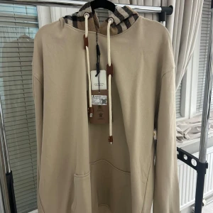 Beige Burberry hoodie med rutmönstrad huva - Säljer en beige hoodie från Burberry med klassisk rutmönstrad huva i brunt, svart, vitt och rött. Tröjan har långa ärmar, snörning med bruna detaljer och en stor magficka. Perfekt för dig som gillar stilrena och lyxiga streetwear-plagg.