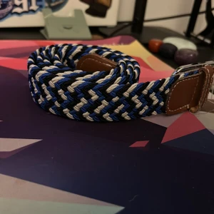 Blå och svart flätat bälte - Säljer ett snyggt flätat bälte med mönster i blått, svart och vitt. Bältet har detaljer i brunt skinn vid spännet och är gjort av ett slitstarkt syntetmaterial. Perfekt för att ge outfiten lite extra edge med sitt coola randiga mönster.