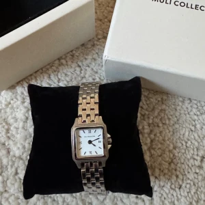 Silverfärgad klocka från Muli Collection - Stilren klocka från Muli Collection med fyrkantig urtavla i vitt och romerska siffror. Armbandet är i silverfärgad metall med klassisk länkdesign. Perfekt accessoar för dig som gillar en tidlös och elegant look.