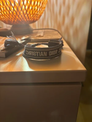 Christian Dior J'Adior blått armband - Snyggt blått tygarmband från Christian Dior med texten 'J'Adior' broderad i vitt. Armbandet har justerbar knytning med tofsar i ändarna och en diskret mönstrad kant. Perfekt accessoar för att lyfta din outfit. Helt nya! Bara skriva vid frågor!